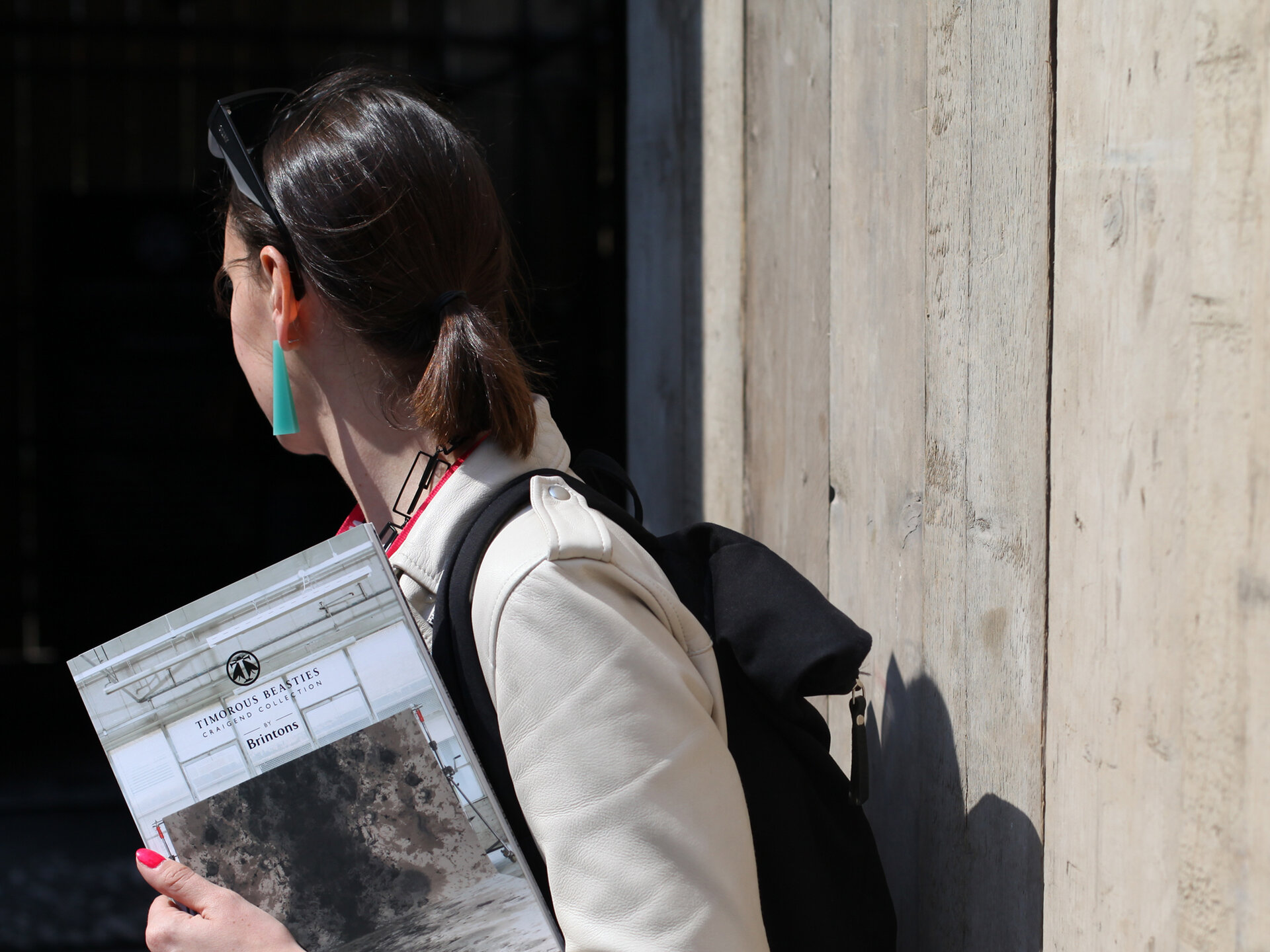A woman turning away from the camera holding a catalogue. She is wearing a beige coat and a black backpack.