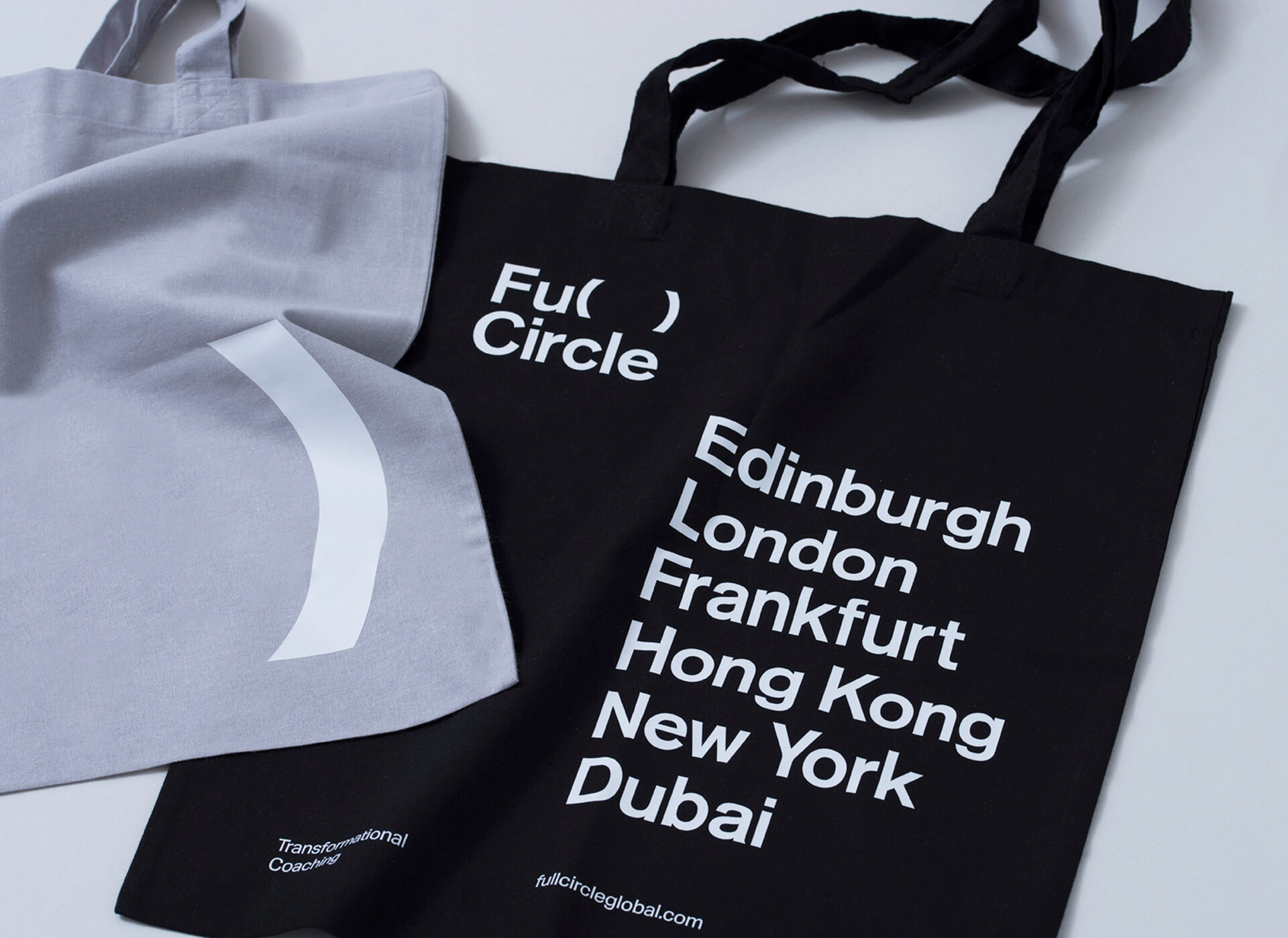 A grey tote bag sits over a black tote bag. The black tote bag has white text on it that reads "Full Circle. Edinburgh,London,Frankfurt,Hong Kong,New York,Dubai".