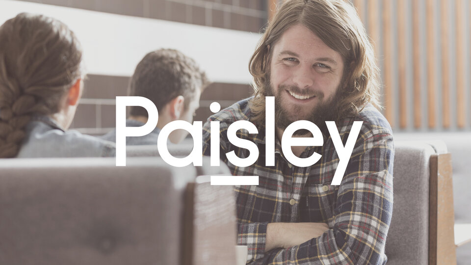 A logo that reads "Paisley" with a line under the "is". Set in white, it sits on a photograph of a man with long hair smiling.