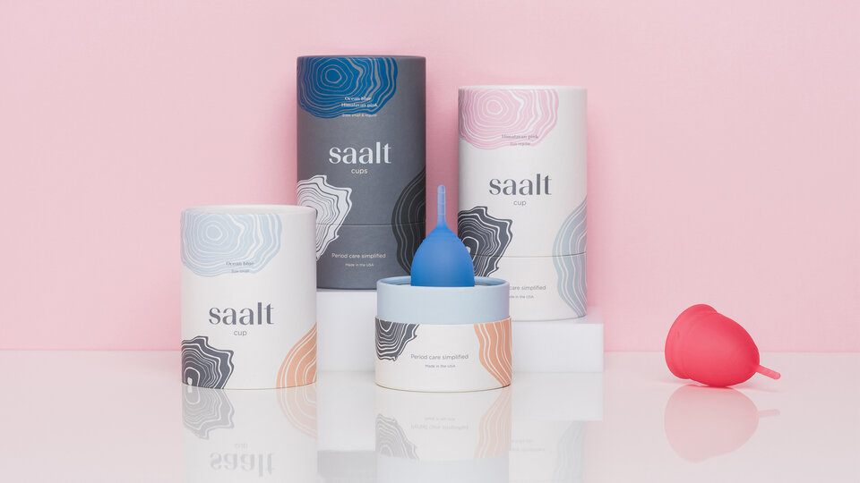 Three Saalt packaging tubes in front of a pink background. There is a blue menstrual cup sitting centrally and a pink one to the right.