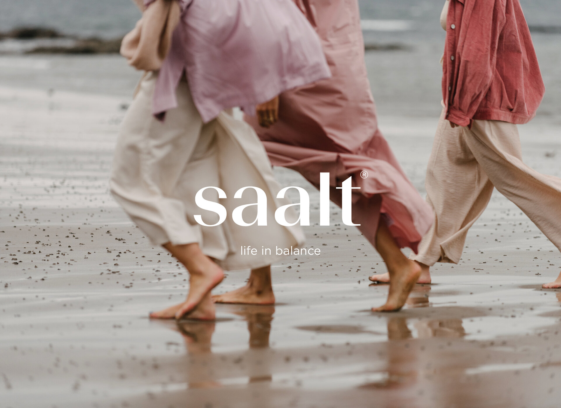 Text that reads "Saalt. Life in balance" over a photograph of three women walking barefoot along a beach. They are all wearing pinky red shades of clothes.