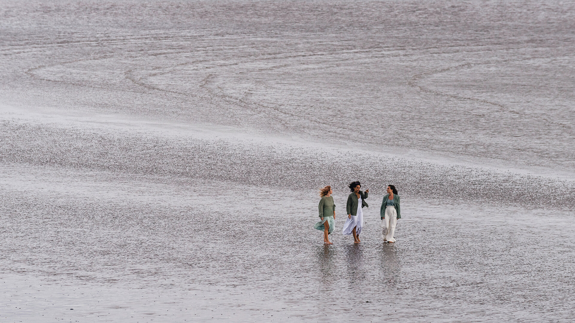 Three women walking beside each other along a beach. There is only sand visible behind them.