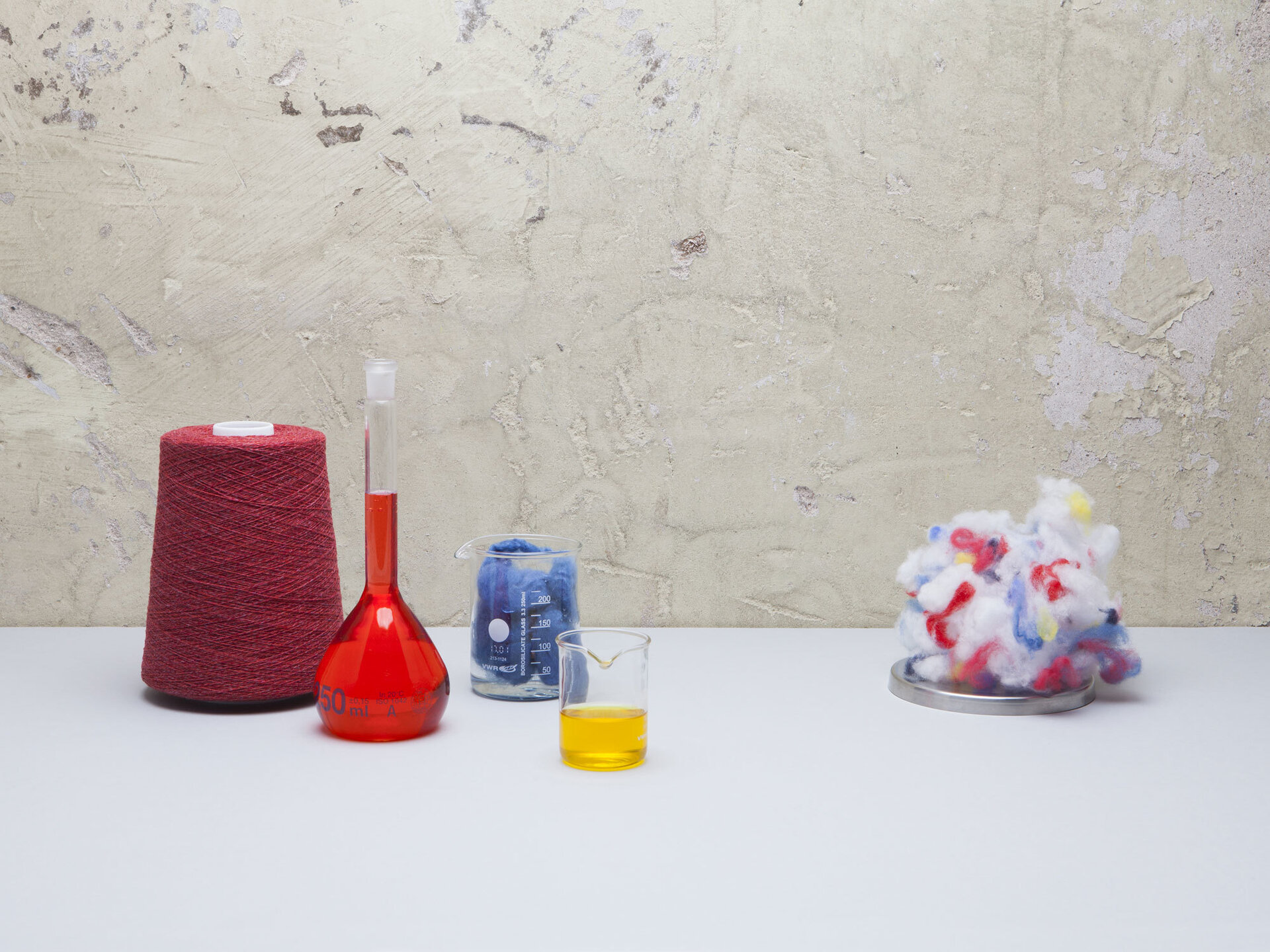 A loom, bottle with red liquid, glass with yellow liquid and a ball of multicoloured fabric sitting on a white tabletop.