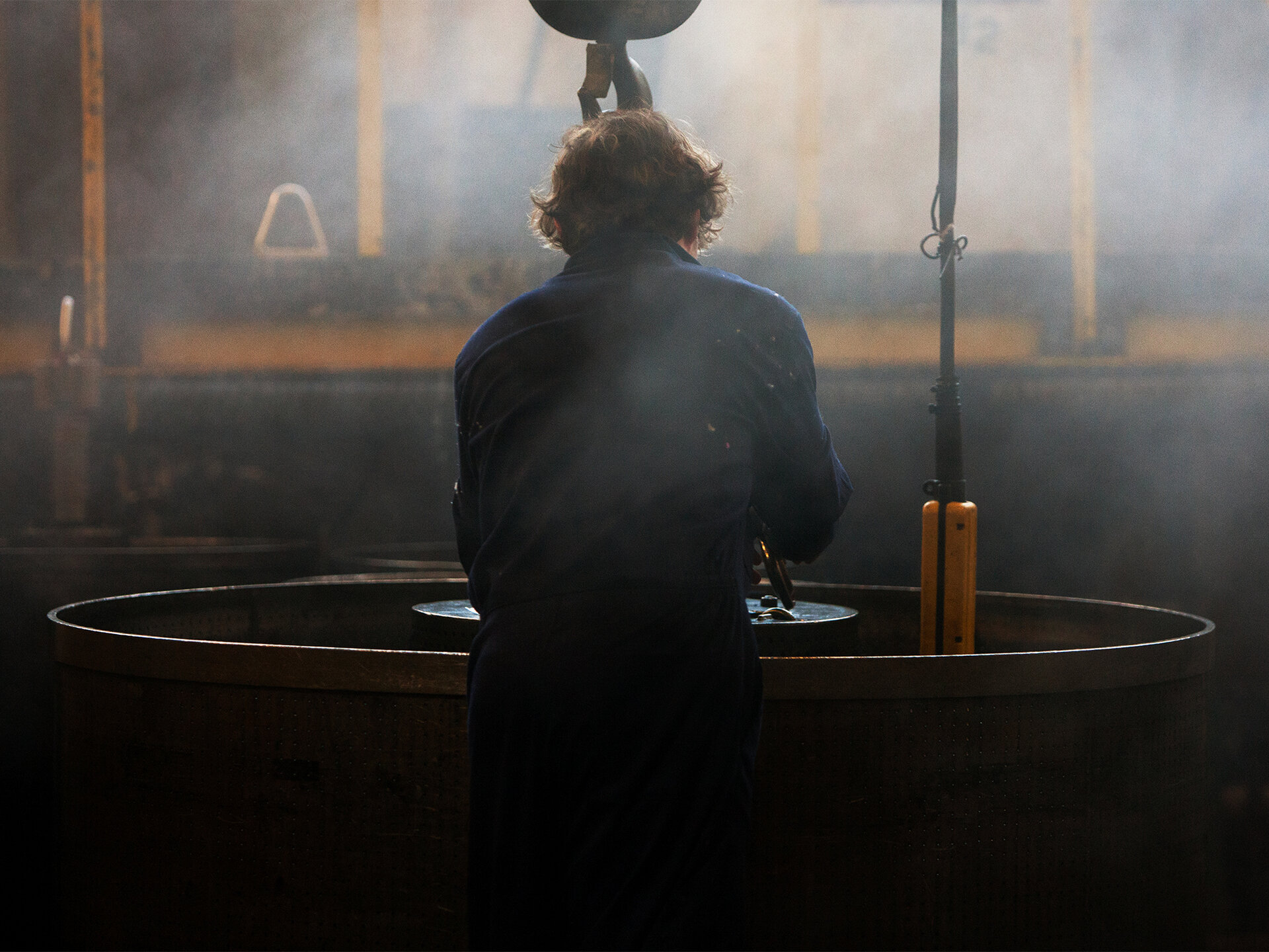 A man standing in the centre in front of a large vat. He is lowering something into it.